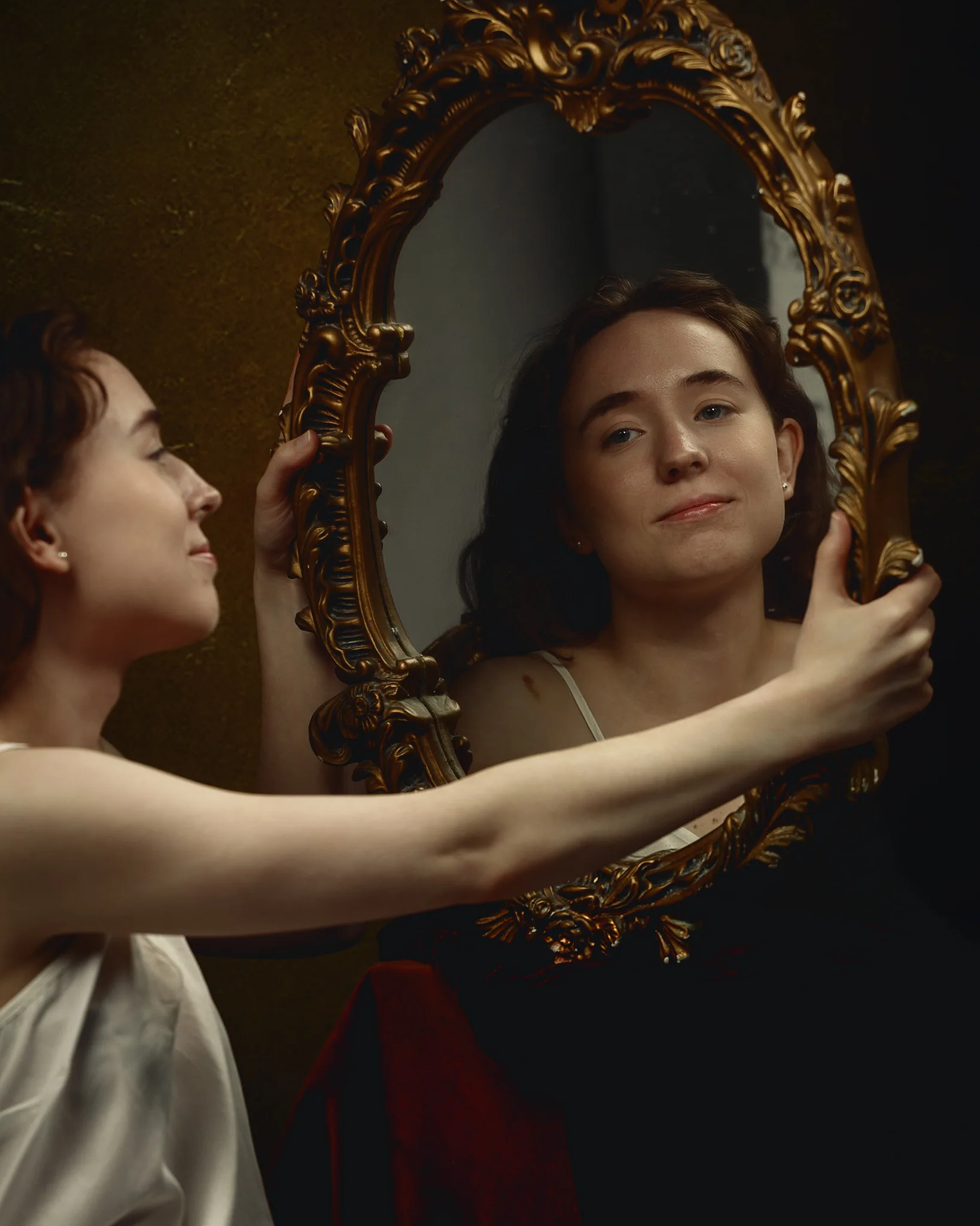 A woman in a white top holds an ornate gold-framed mirror, gazing at her reflection against a dark background.