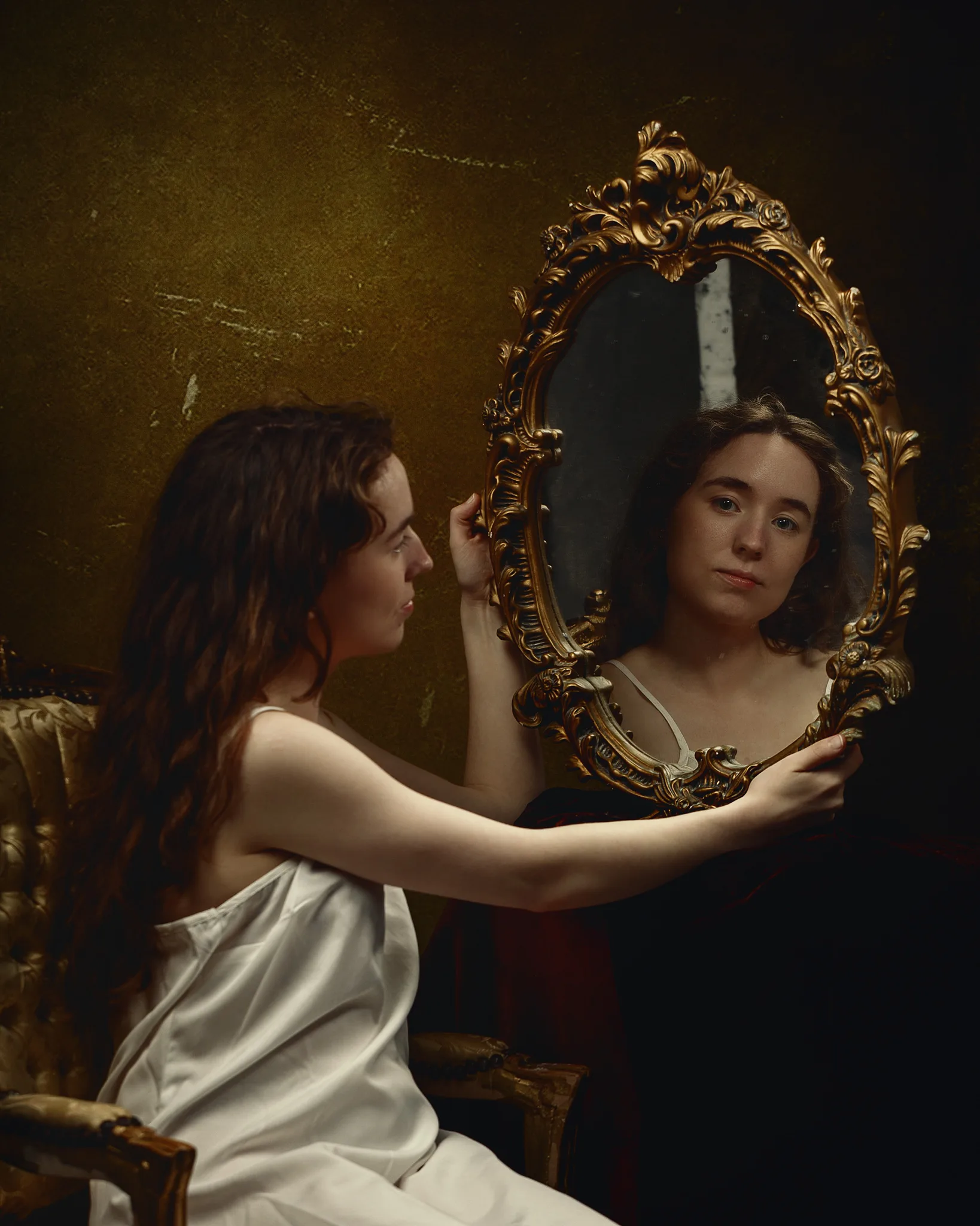A woman in a white garment holds an ornate gold-framed mirror, gazing at her reflection against a textured golden-brown wall.