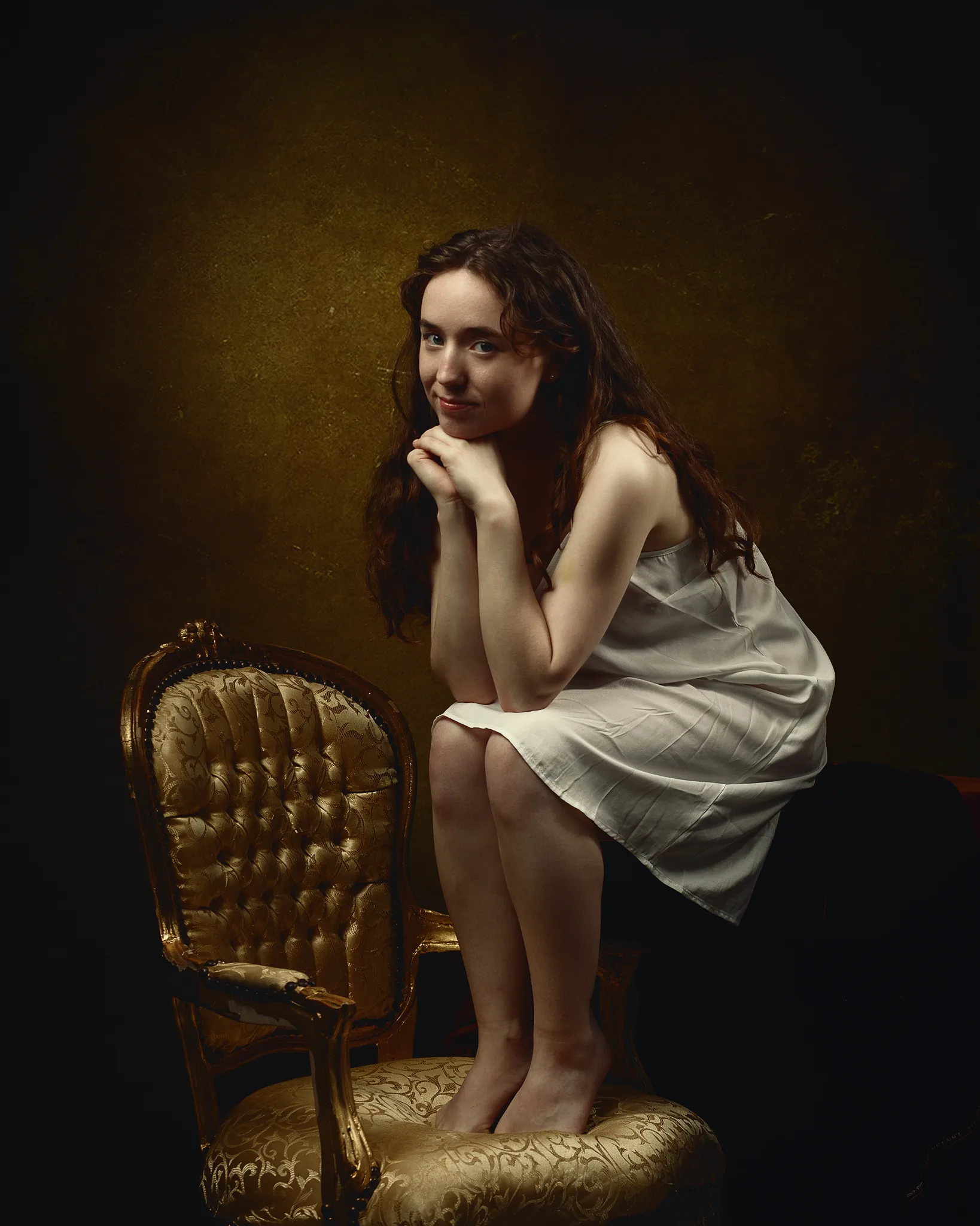 A woman with long dark hair sits on an ornate gold chair, wearing a white slip dress, against a dark golden-brown background.