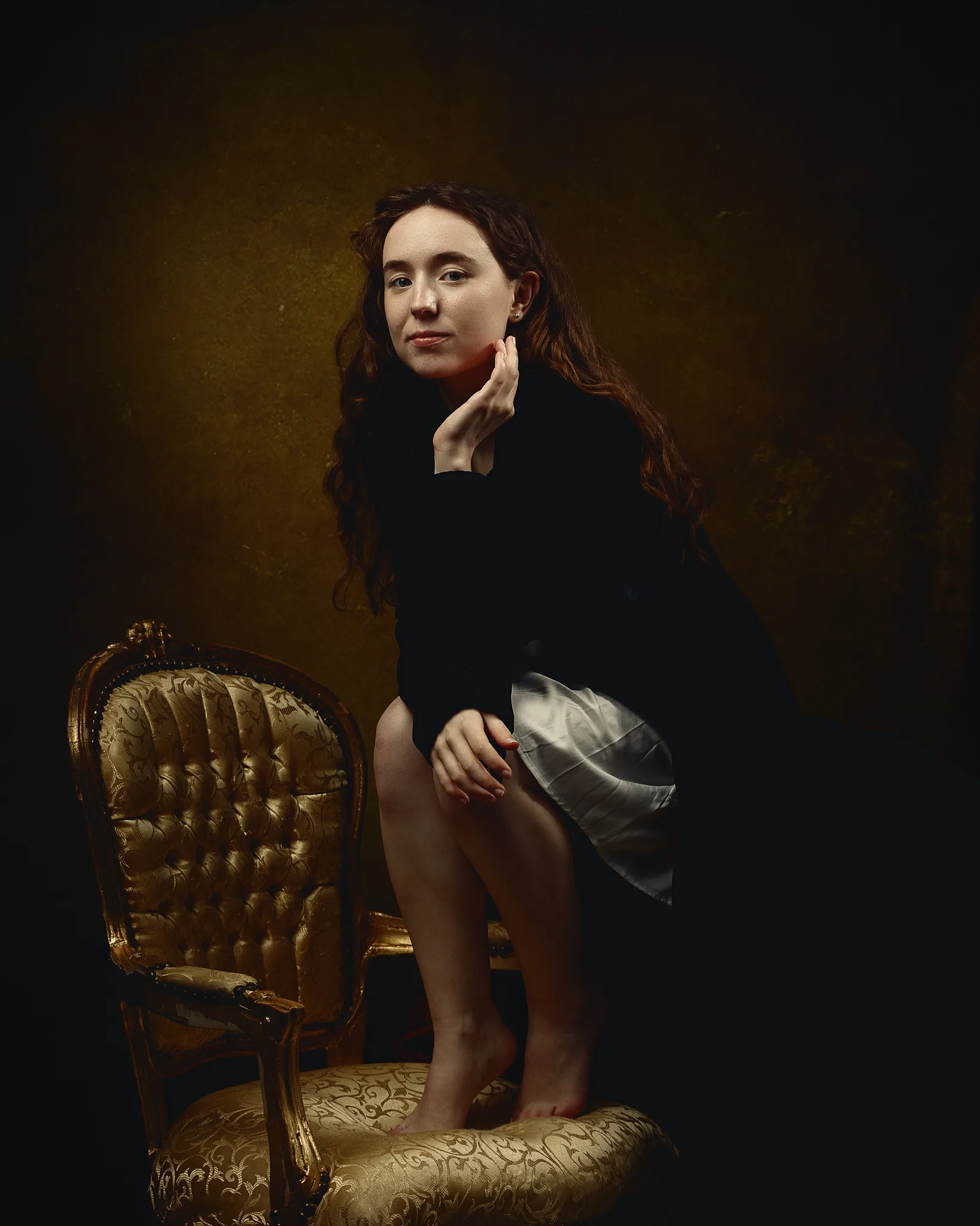 A woman with long reddish-brown hair stands beside an ornate gold chair against a dark background, touching her face thoughtfully.