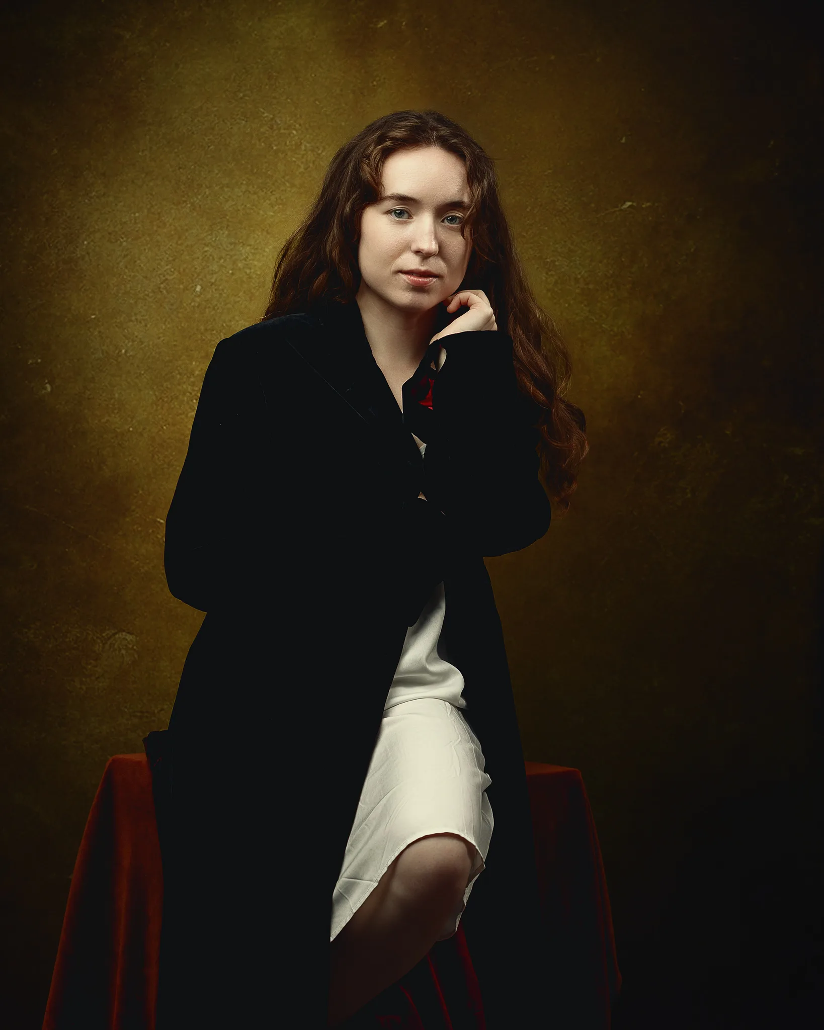 A woman with long dark hair sits on a red chair wearing a black blazer and white skirt against a golden-brown backdrop.