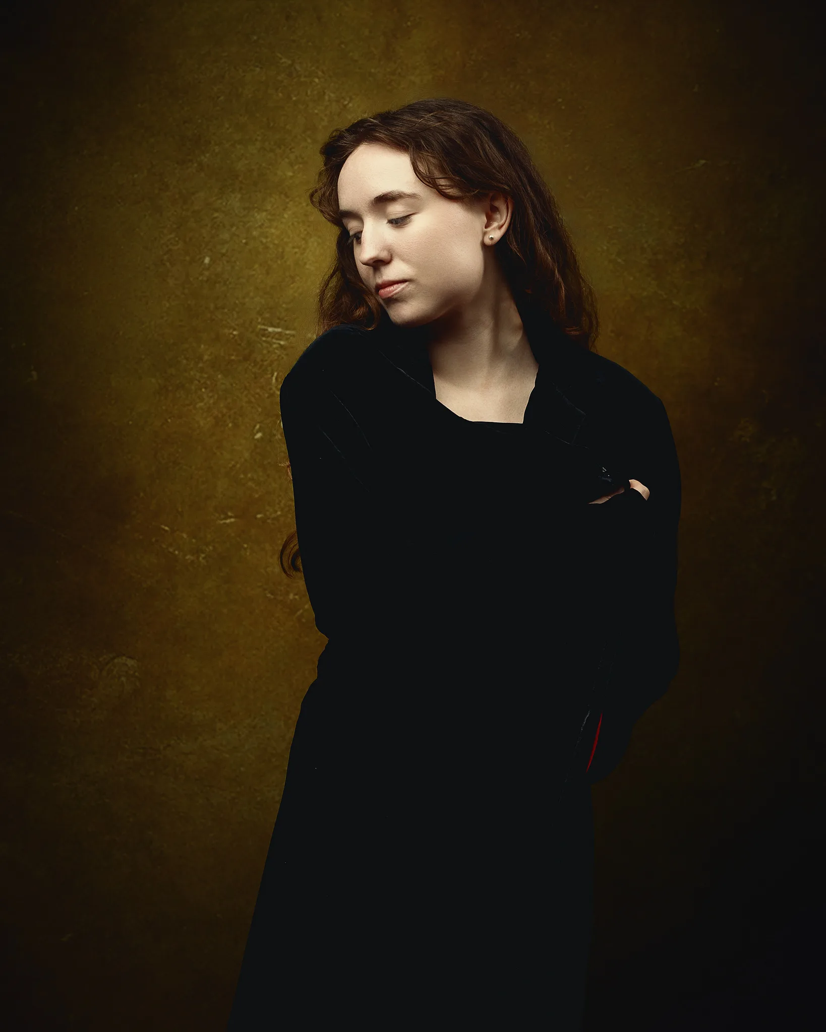 A young woman with long dark hair wearing a black coat stands against a textured gold-brown background, looking downward.