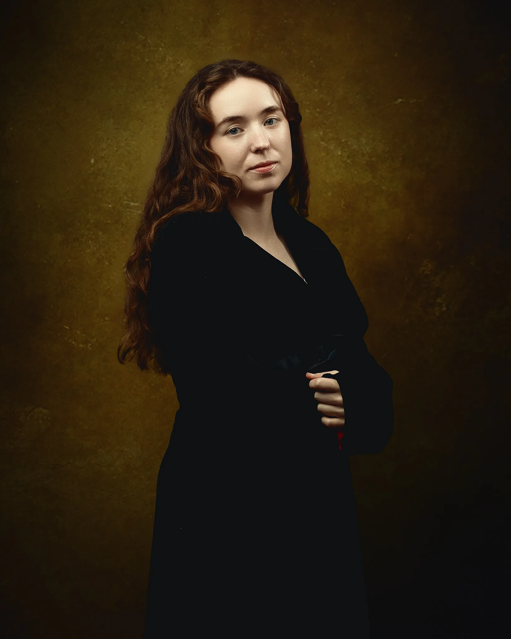 A young woman with long wavy brown hair wearing a black coat stands against a muted gold-brown background, looking toward the camera.