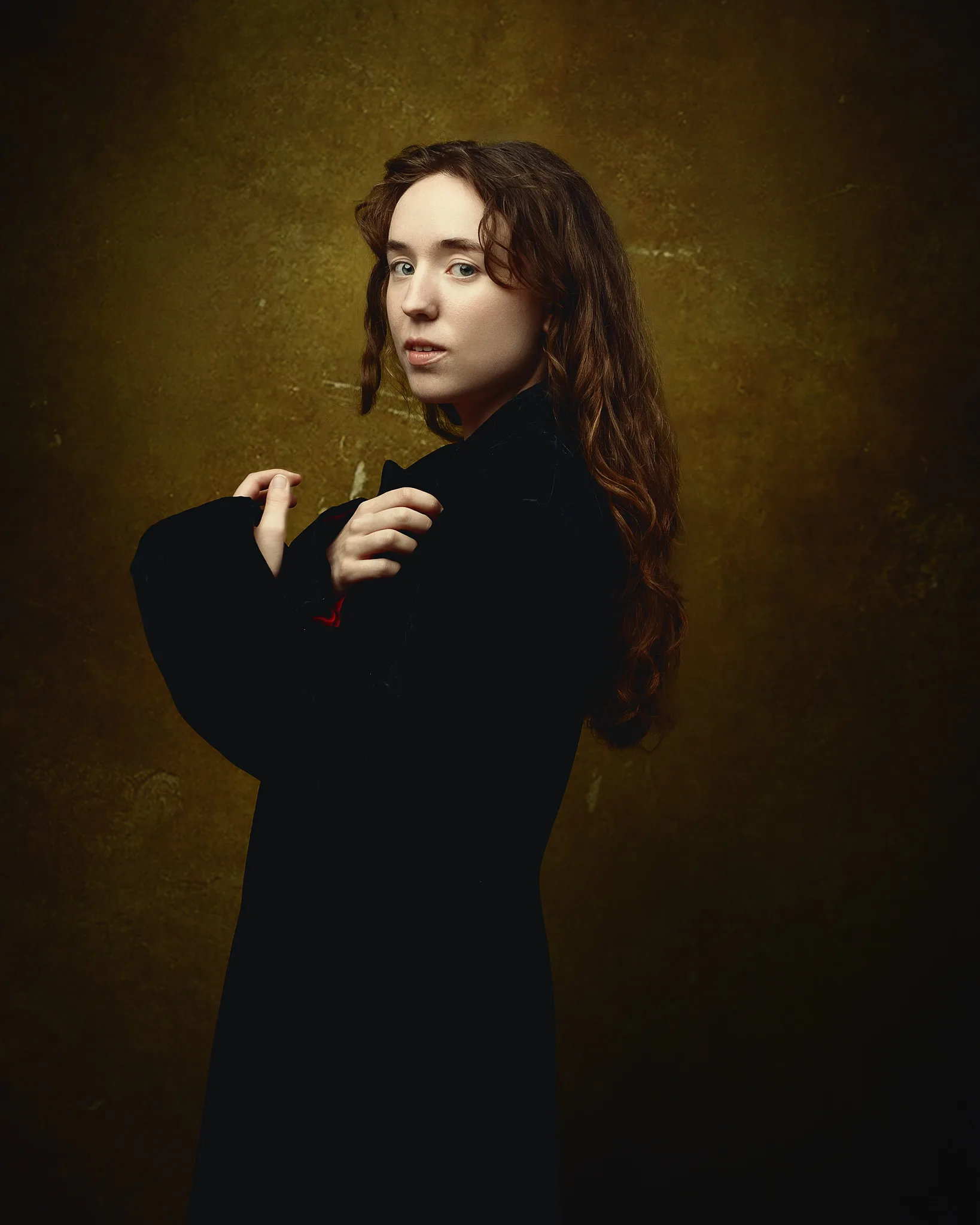 A person with long brown hair wearing a black blazer stands against a mottled gold-brown background, looking over their shoulder.