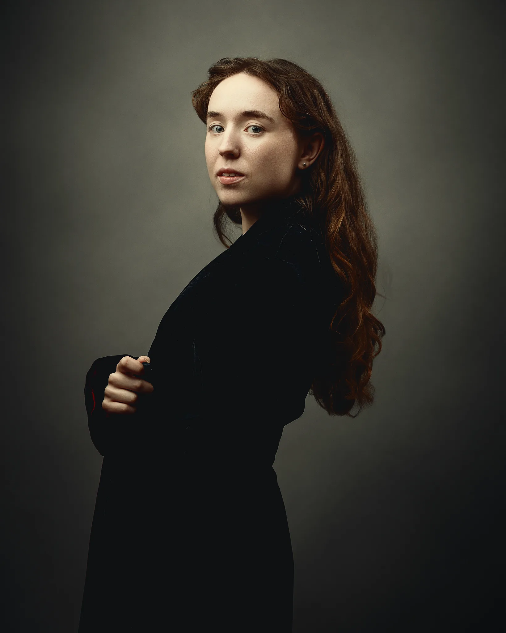 Woman with long reddish-brown hair wearing a black blazer, looking over her shoulder against a neutral grey background.