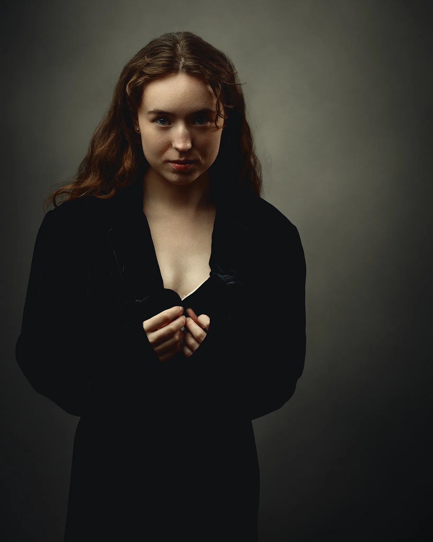 A young woman with long auburn hair wearing a black cardigan stands against a grey background, looking downward with hands clasped.