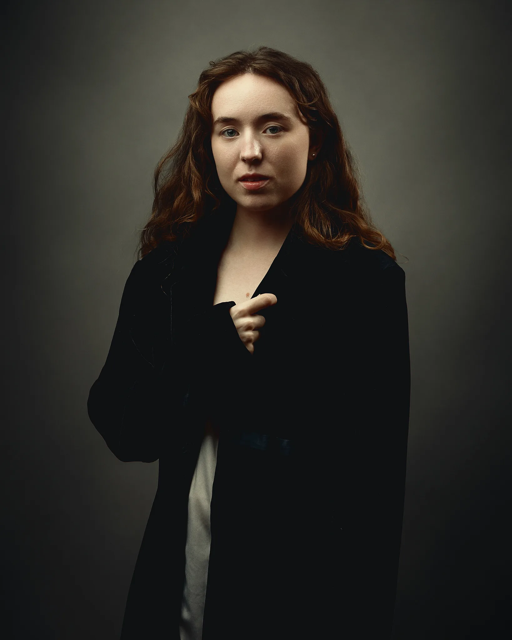 A woman with reddish-brown hair wearing a black blazer over a white top, photographed against a neutral grey background.