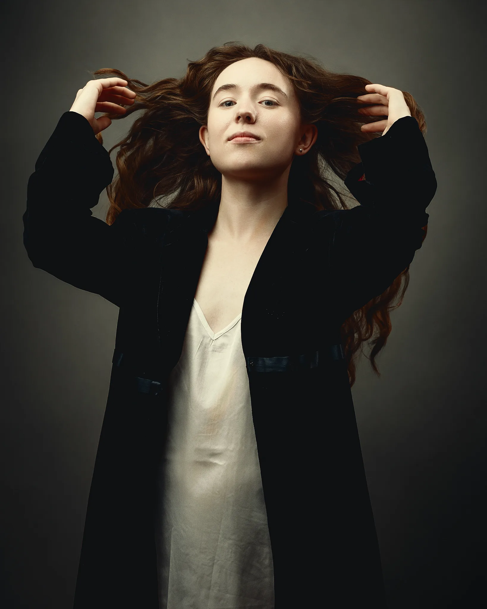 A person with long brown hair wearing a black blazer and cream slip dress, hands raised to their hair, against a neutral grey background.