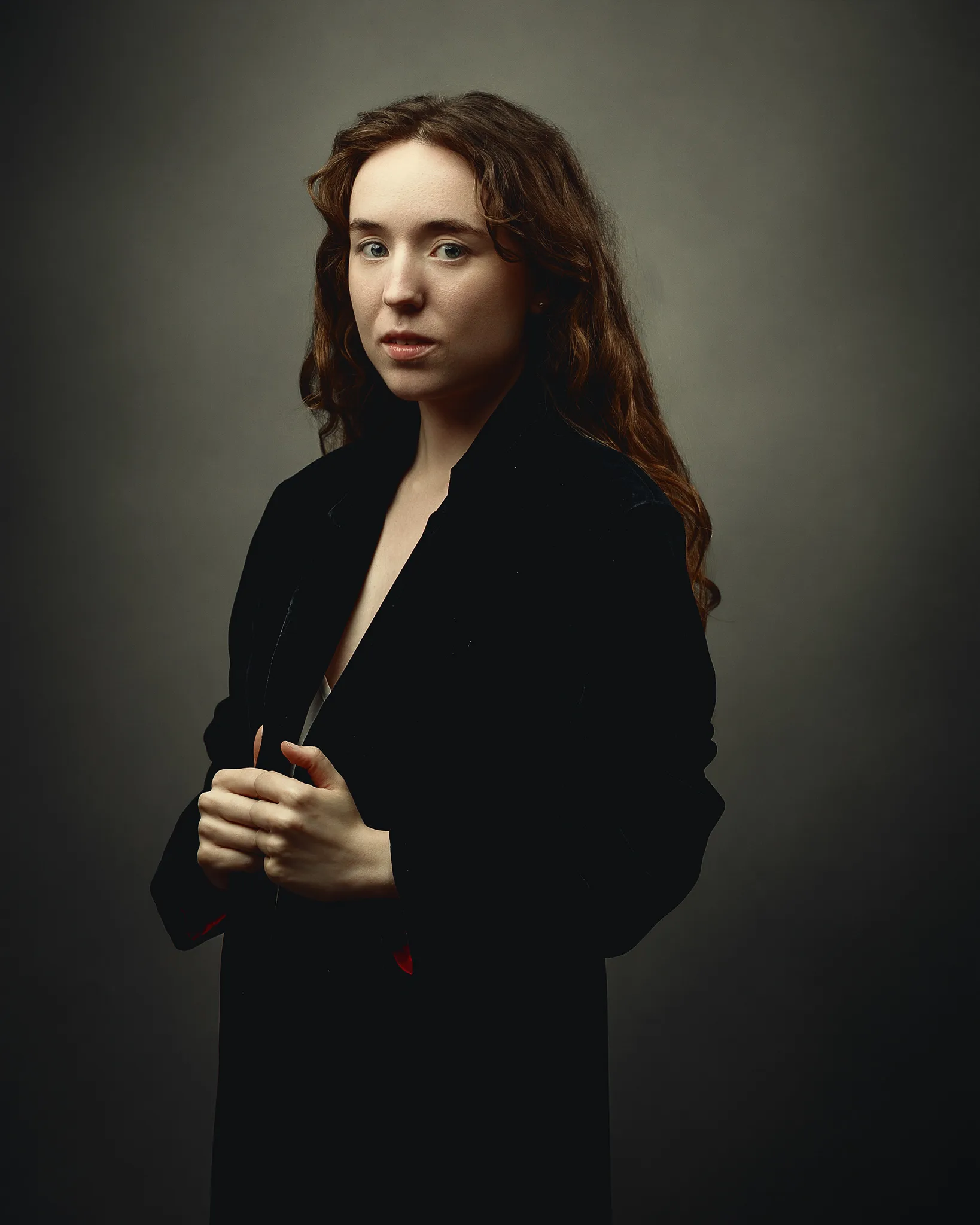 A woman with long reddish-brown hair wearing a black blazer stands against a neutral grey background, gazing to the side.