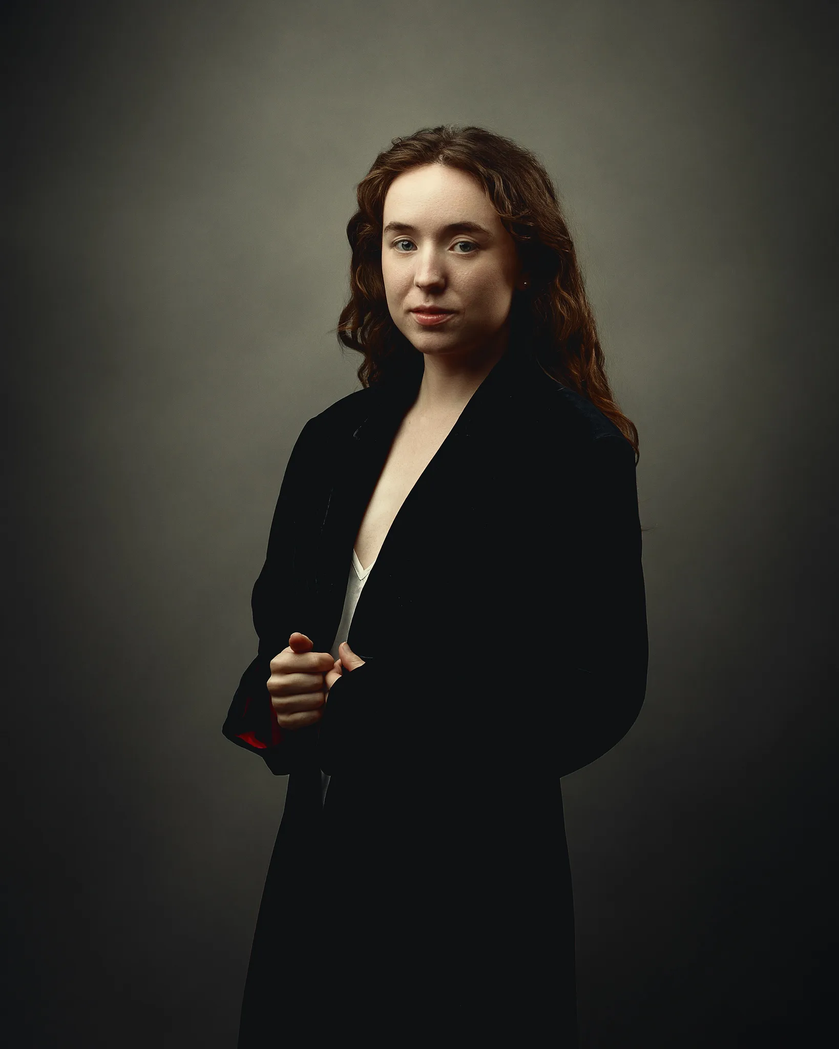 A woman with long reddish-brown wavy hair wears a black blazer over a white shirt against a neutral grey background.