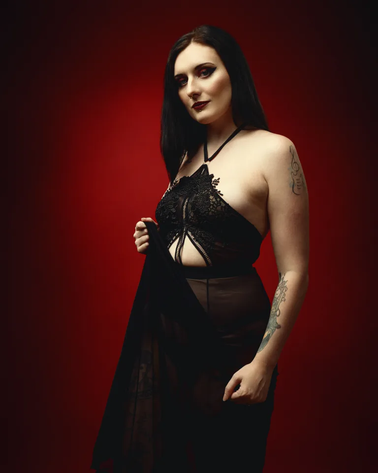 A woman with long dark hair and dark makeup stands against a red background, wearing a black lace halter top and sheer skirt.