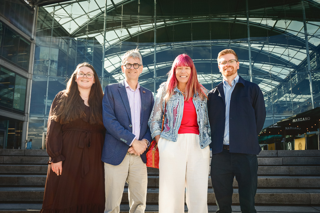 Four people stand together smiling in front of a modern glass building with a steel framework roof.