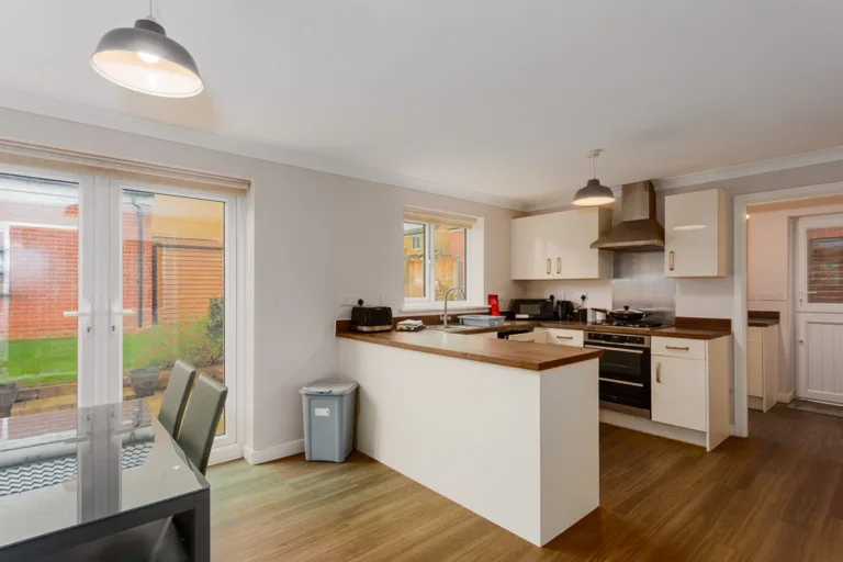 A modern kitchen with white cabinets, wooden worktops, and a central island. Pendant lights hang from the ceiling. French doors lead to a garden.