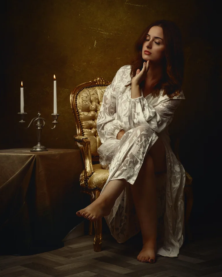 Woman in white robe sitting on gilded chair beside lit candelabra against dark gold background.