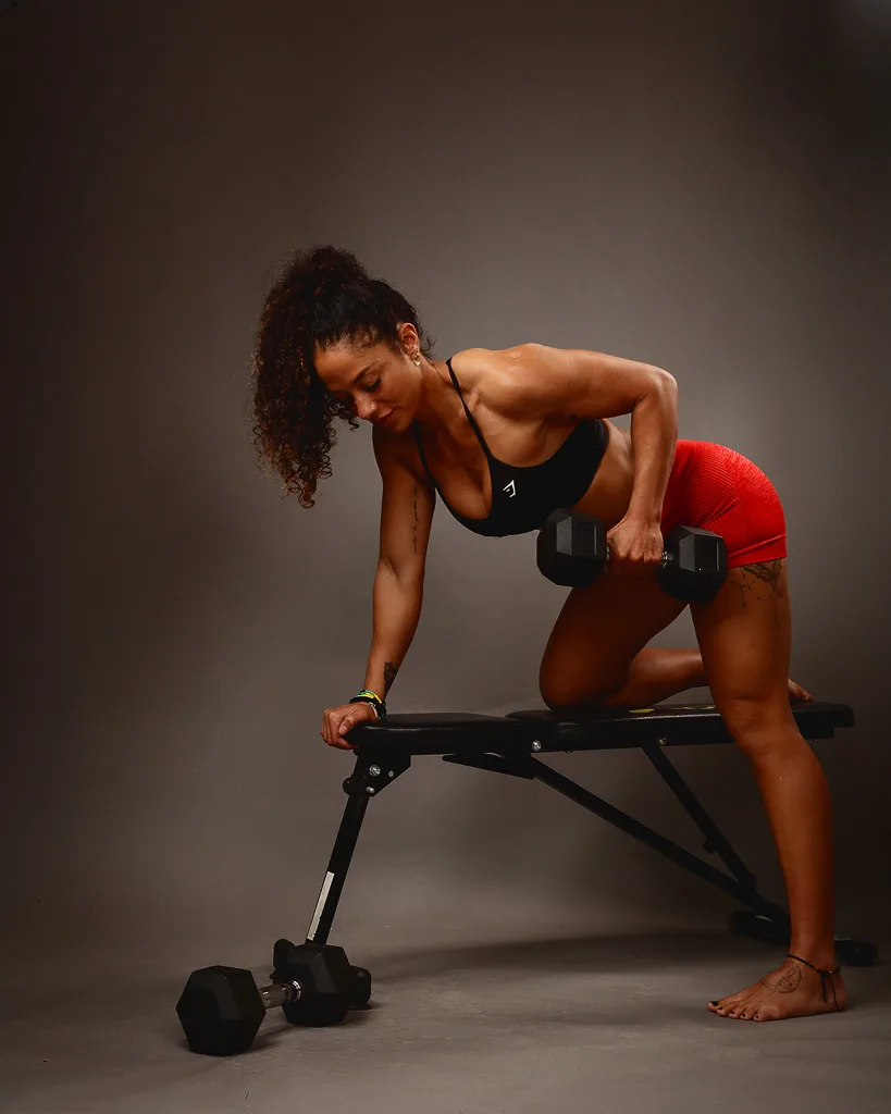A fit woman performs a dumbbell row exercise on a weight bench against a neutral grey background.