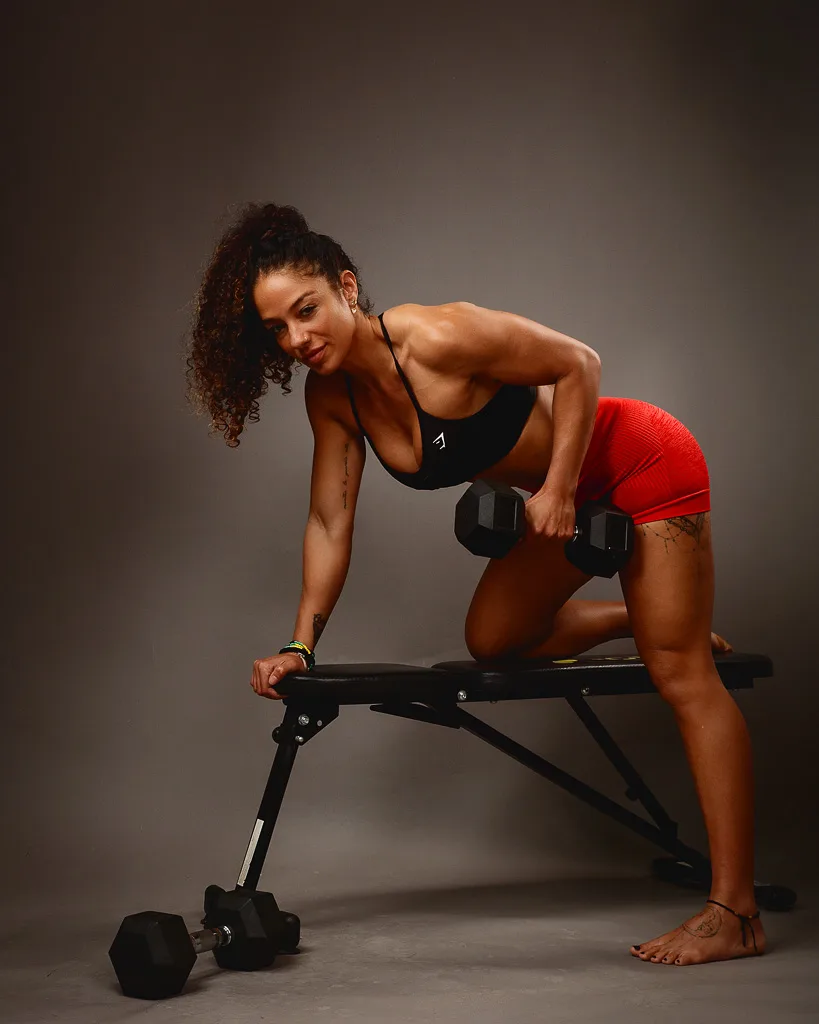 Athletic woman in black sports top and red shorts holding dumbbells while kneeling on a weight bench against a grey background.