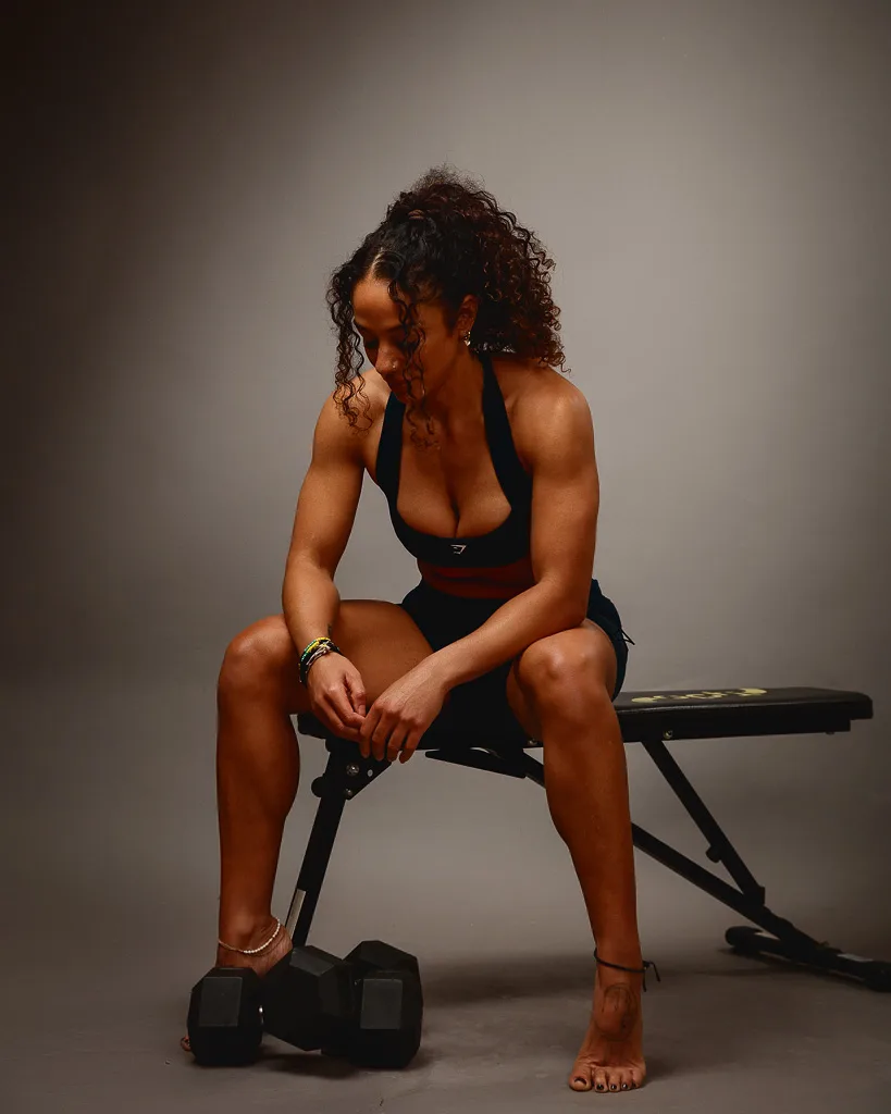 A woman in athletic wear sits on a weight bench with dumbbells nearby against a neutral grey background.