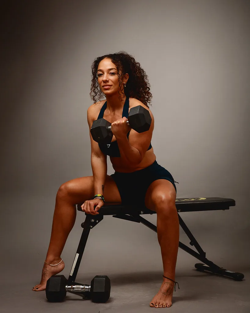 A fit woman in black sportswear holds dumbbells while seated on a weight bench against a neutral grey background.