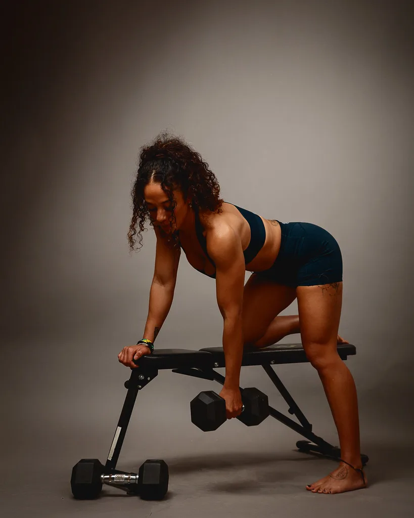 A woman performs a dumbbell exercise on a weight bench against a neutral grey background.