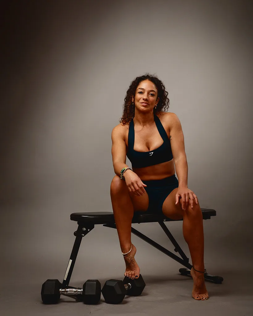 A woman in black fitness attire sits on a weight bench with dumbbells on the floor beside her against a neutral grey background.