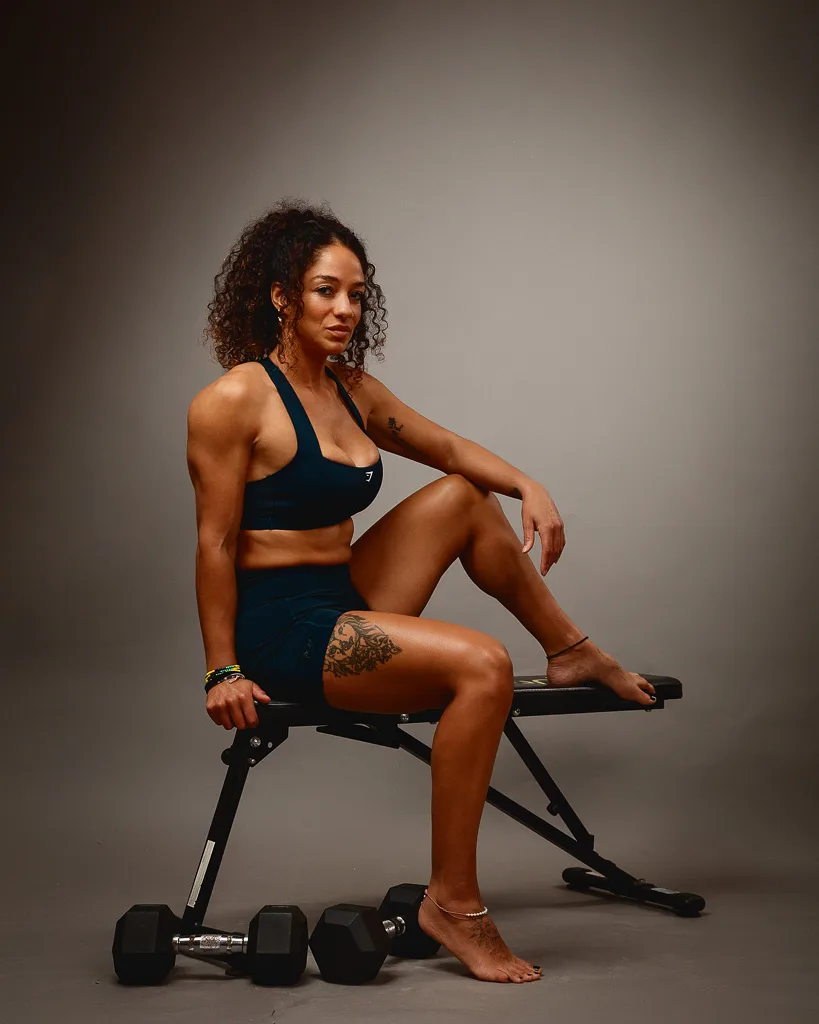 A woman in dark athletic wear sits on a weight bench with dumbbells beside her against a neutral grey background.
