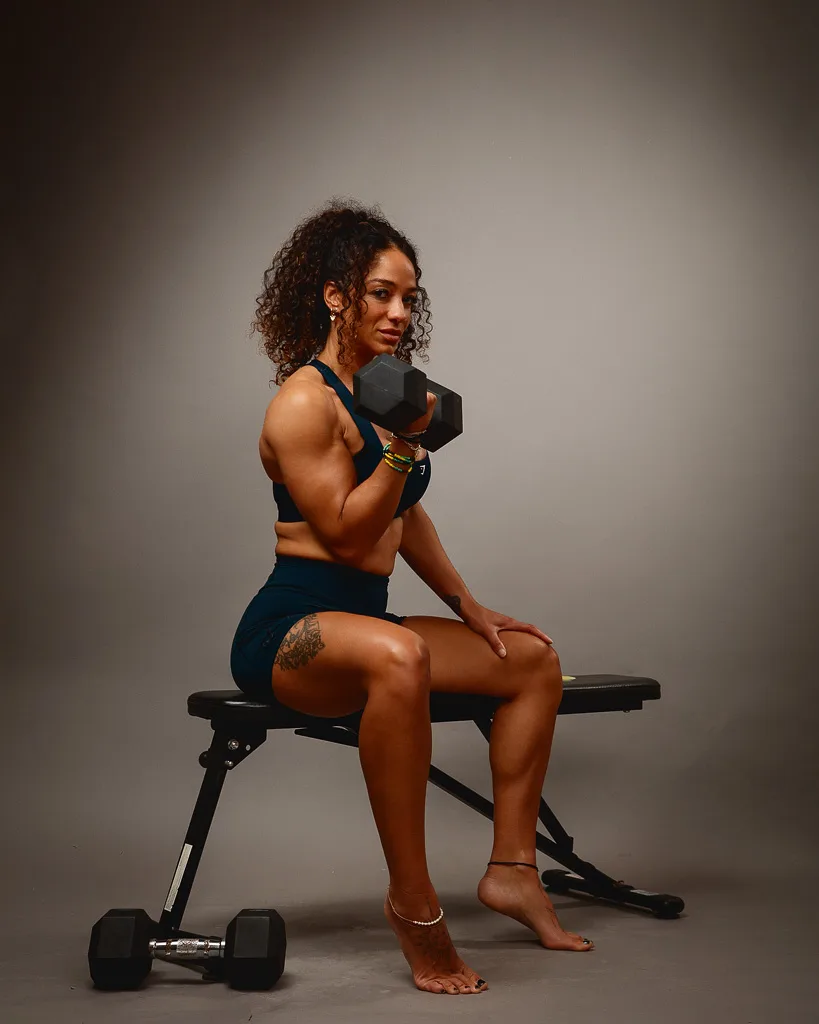 Woman in fitness attire holding a dumbbell, seated on a weight bench with dumbbells beside her against a neutral background.