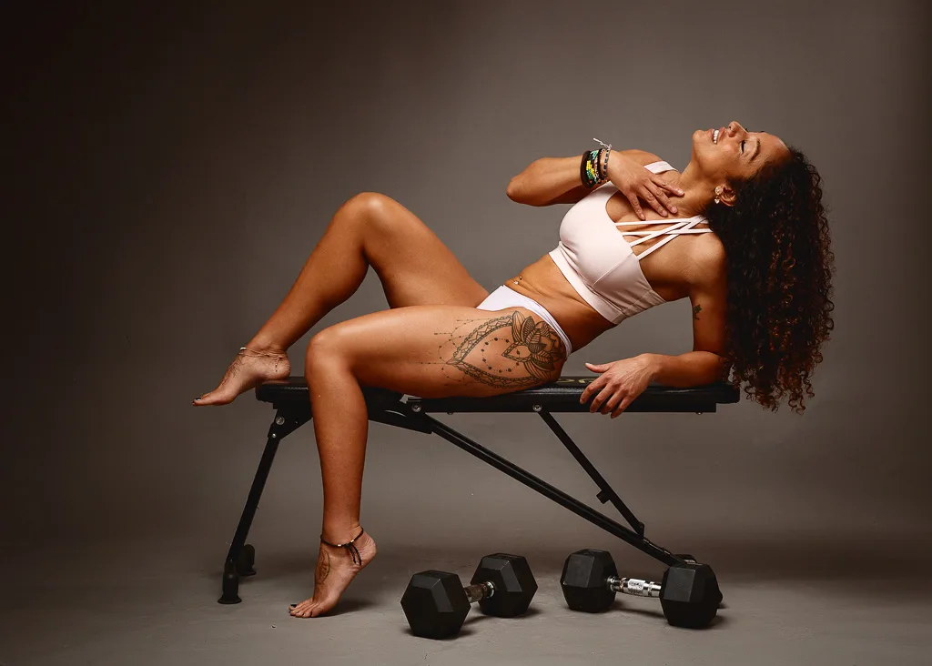 A woman reclines on a weight bench in athletic wear, surrounded by dumbbells against a neutral gray background.