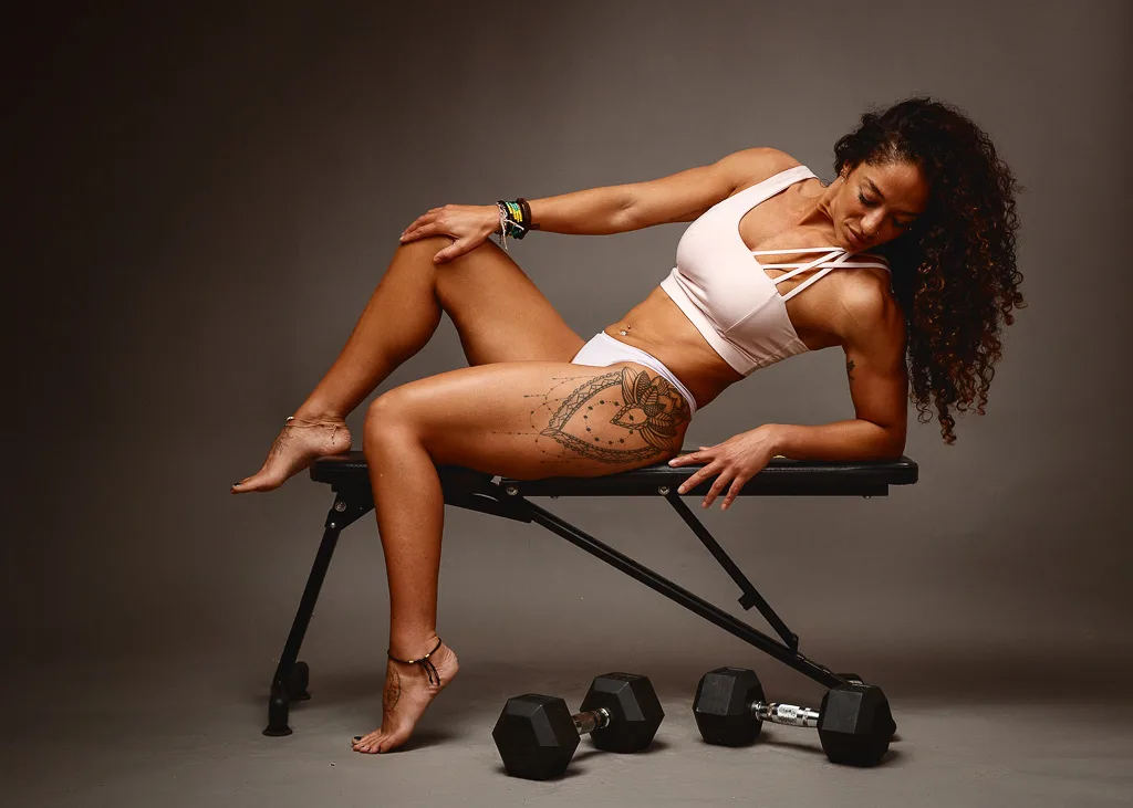 Woman in white athletic wear reclining on a weight bench with dumbbells on the floor against a grey background.