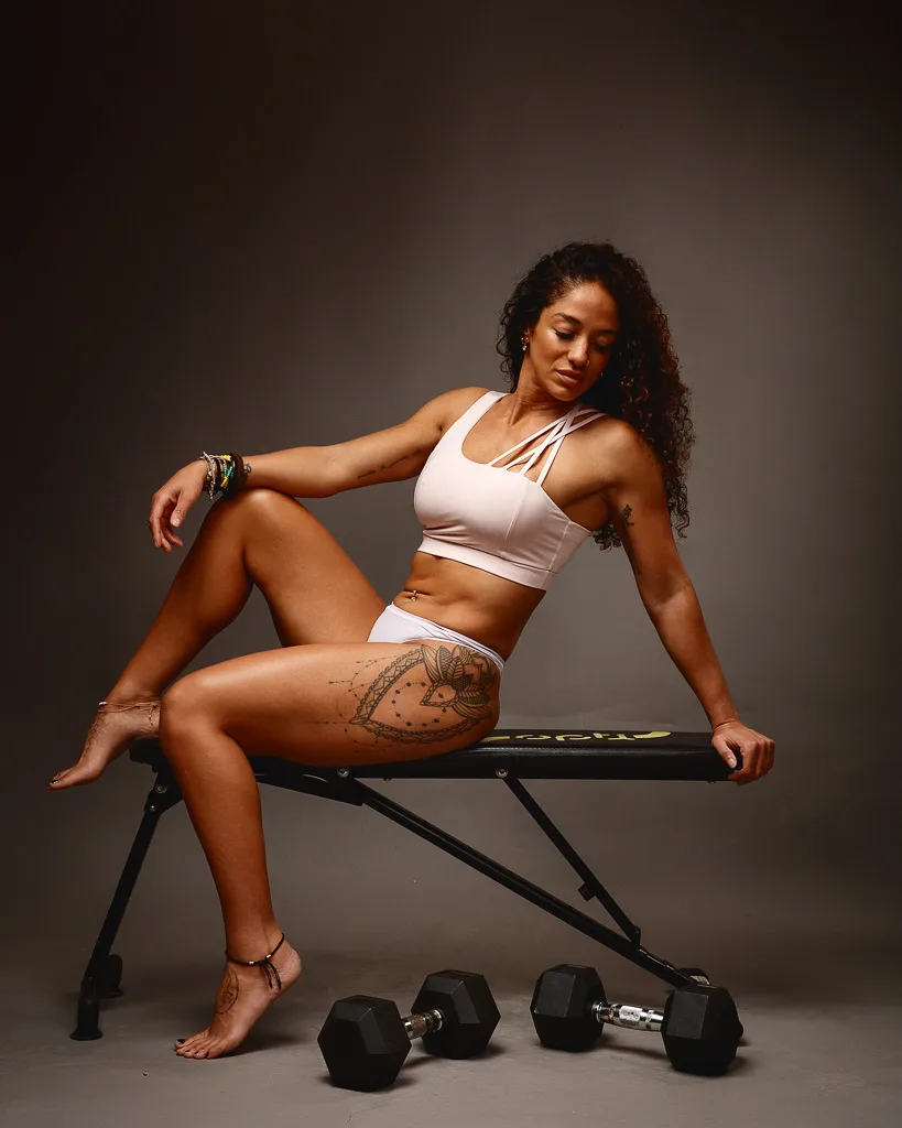 A woman in white fitness wear sits on a bench with dumbbells on the floor beside her, against a neutral grey background.