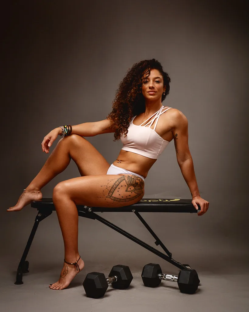 A woman in white athletic wear sits on a weight bench with dumbbells on the floor beside her against a grey background.
