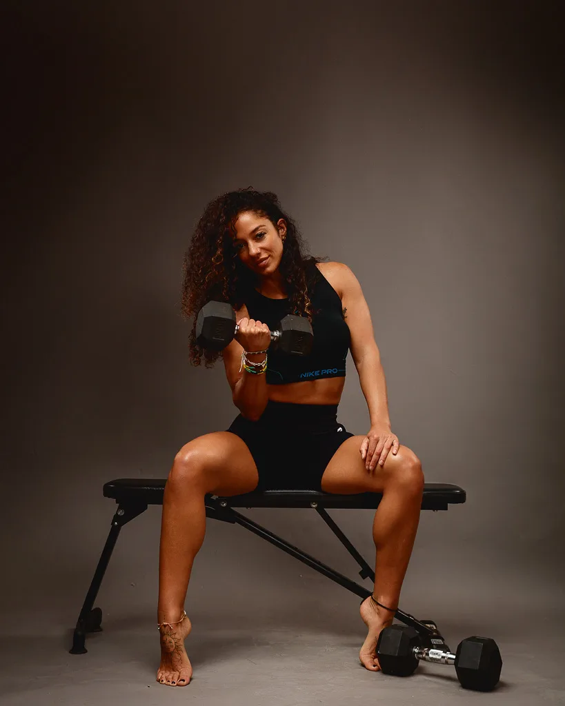 A woman in black athletic wear sits on a weight bench holding a dumbbell, with another dumbbell on the floor beside her.
