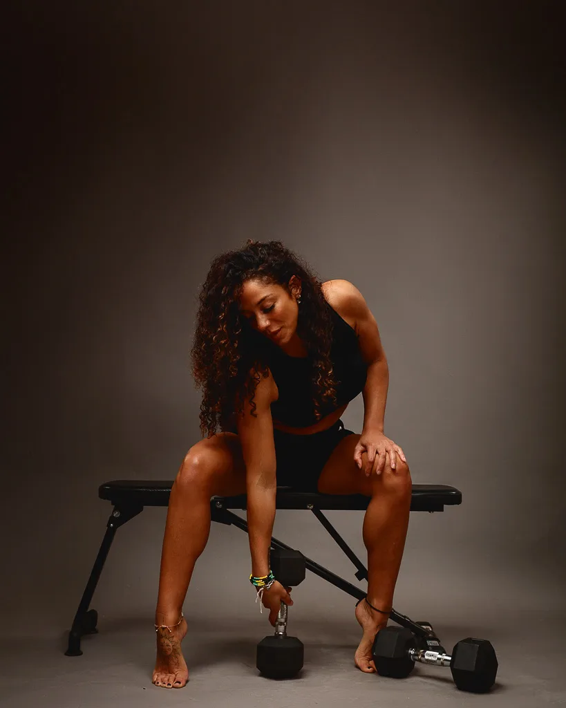 A woman in black workout attire sits on a weight bench, holding a dumbbell, with additional weights on the floor beside her.