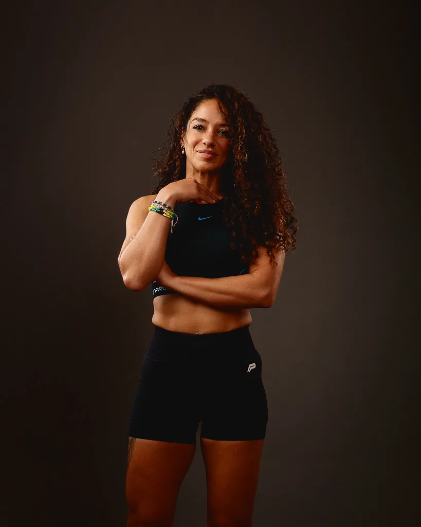 A woman in athletic wear stands against a brown background with arms crossed, wearing a black Nike crop top and black shorts.