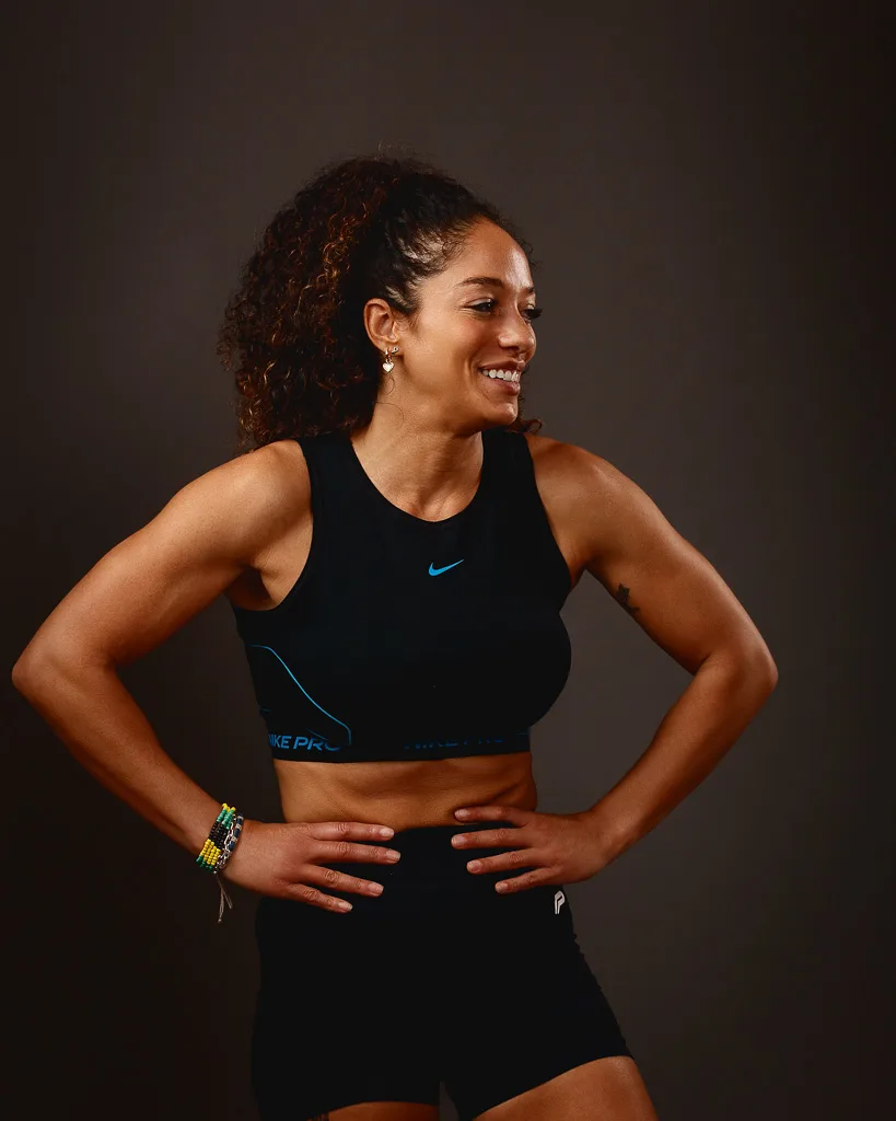 A woman in Nike sportswear poses confidently against a brown background, smiling to the side with hands on hips.