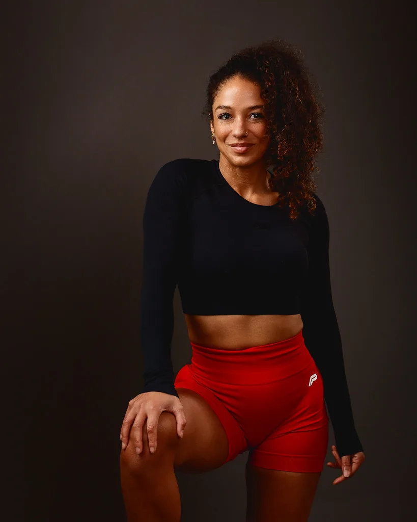 A woman in athletic wear poses against a dark background, wearing a black crop top and red shorts.