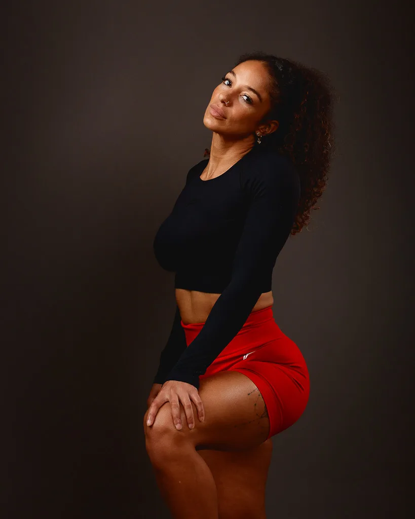 A woman in a black long-sleeved crop top and red shorts poses against a dark background, looking upward over her shoulder.