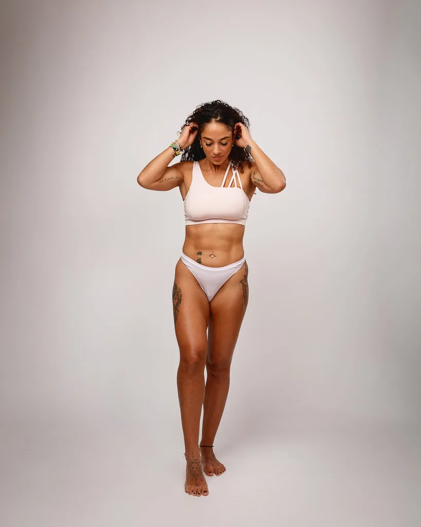 A woman in white sports bikini and crop top stands against a plain grey background, hands raised to her head.
