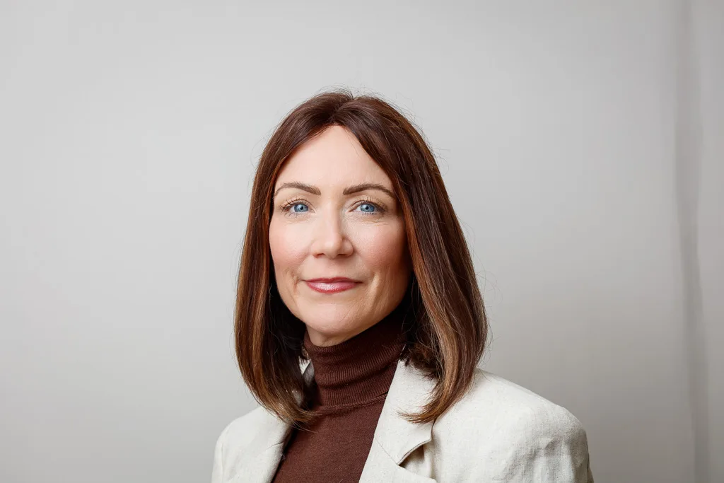 A woman with shoulder-length auburn hair and blue eyes, wearing a brown turtleneck and cream blazer, smiles at the camera against a plain grey background.
