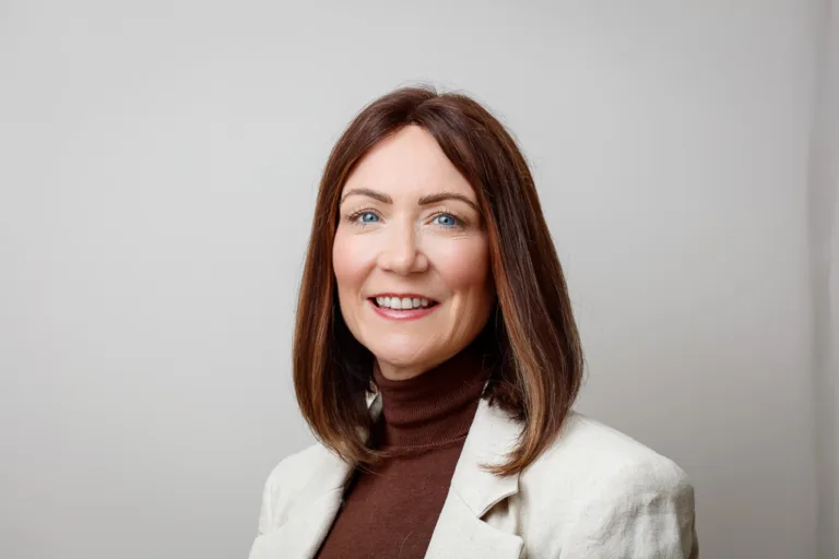 A woman with shoulder-length brown hair smiles at the camera. She wears a burgundy turtleneck and cream blazer against a plain grey background.