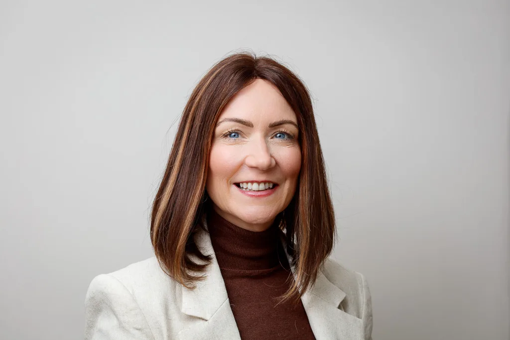 A woman with shoulder-length brown hair smiles at the camera. She wears a dark turtleneck and light blazer against a plain grey background.