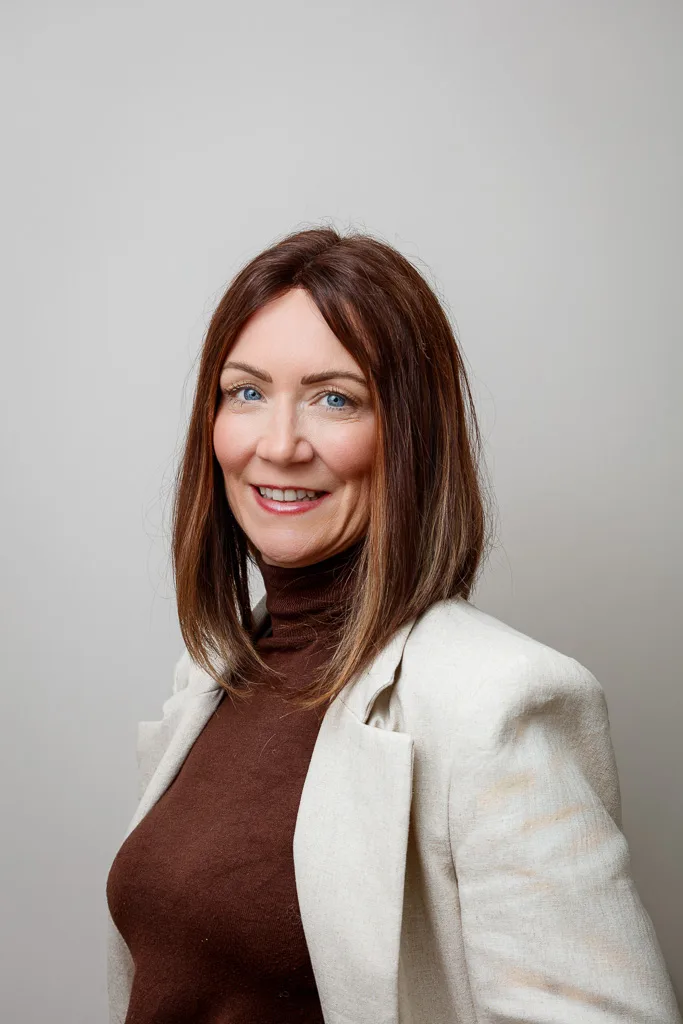 A woman with shoulder-length auburn hair smiles at the camera, wearing a burgundy turtleneck and cream blazer against a plain grey background.