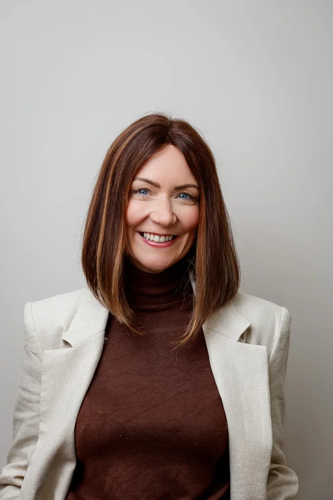 A woman with shoulder-length auburn hair smiles at the camera, wearing a brown turtleneck and cream blazer against a grey background.
