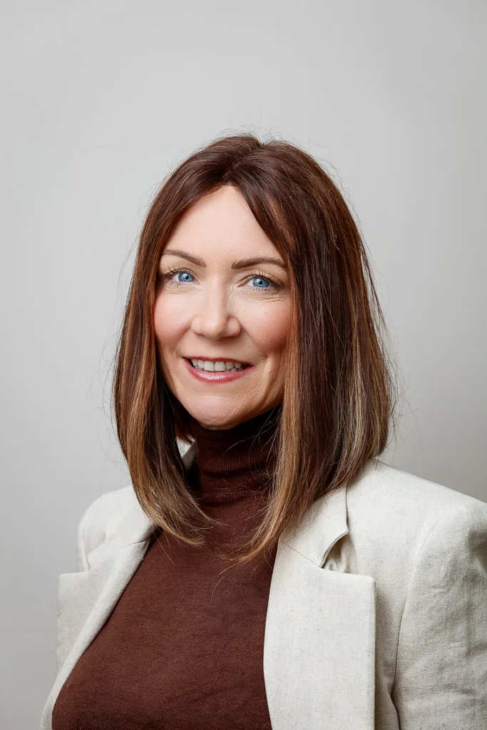A woman with shoulder-length auburn hair and blue eyes smiles at the camera, wearing a brown turtleneck and cream blazer against a grey background.
