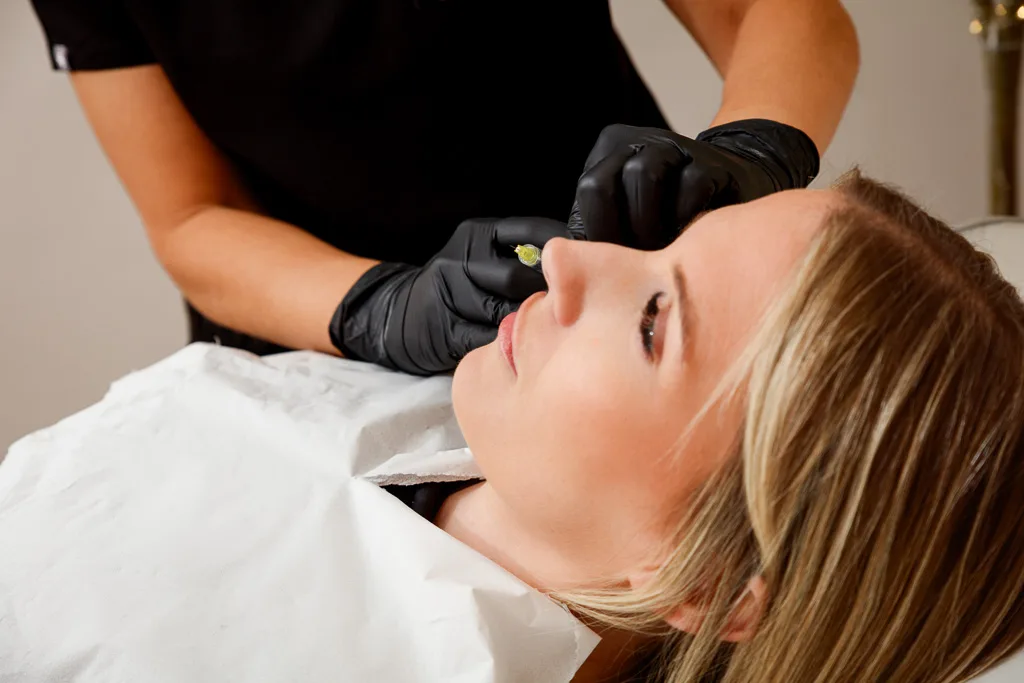 A practitioner wearing black gloves performs a facial treatment on a relaxed client lying on a white bed.