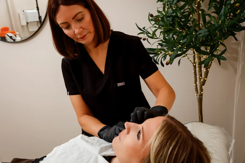 A healthcare professional in black scrubs and gloves examines a patient's face in a clinical treatment room with potted plants.