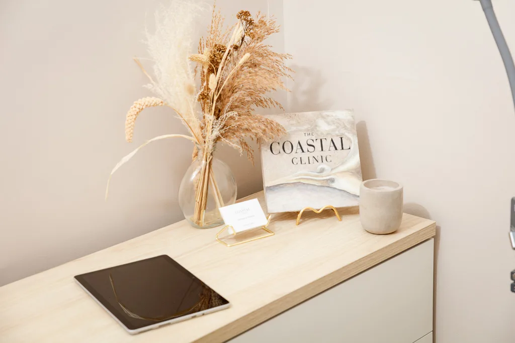 A minimalist reception desk displaying dried pampas grass, a book titled The Coastal Clinic, a small candle, and a tablet device.