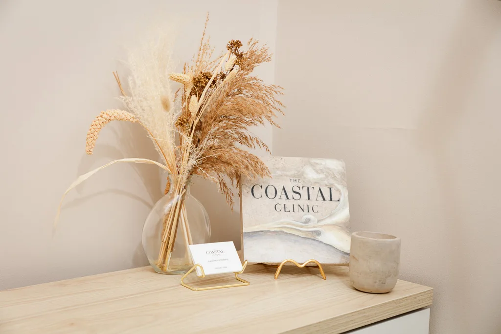 A desk display featuring a vase with dried pampas grass, a sign reading The Coastal Clinic, and a concrete candle.