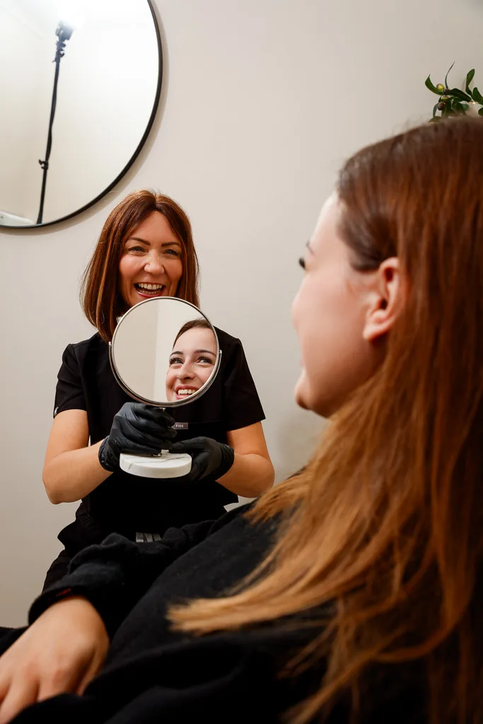 A beautician holds a hand mirror toward a smiling client with reddish-brown hair during a salon consultation.
