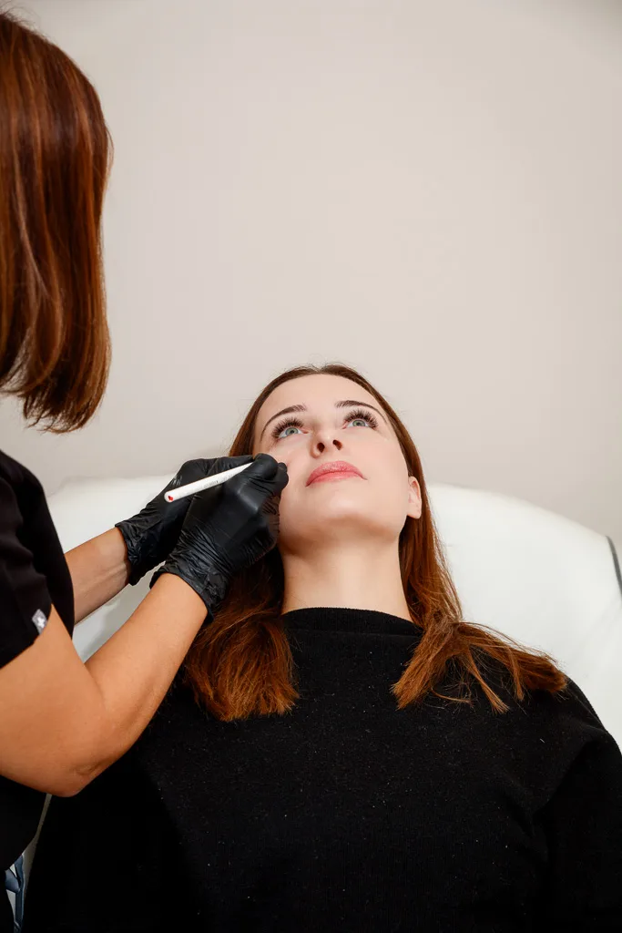 A practitioner in black gloves examines a woman's face with a dermatology tool against a white background.