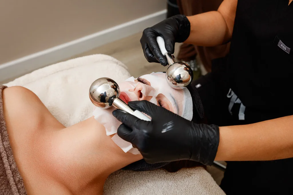 A therapist in black gloves uses metal facial massage balls on a client's face during a beauty treatment session.